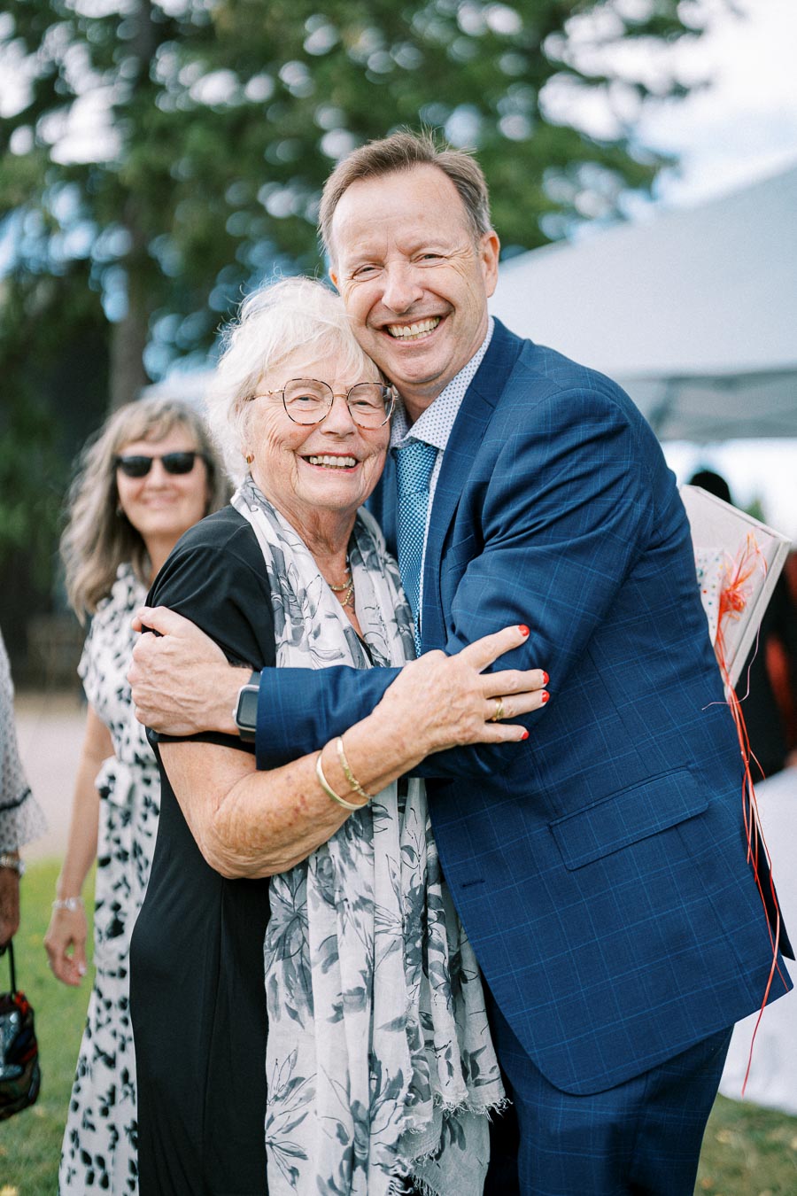 A joyful elderly woman with glasses and a floral scarf hugging a man in a blue suit, both smiling warmly at an outdoor event.