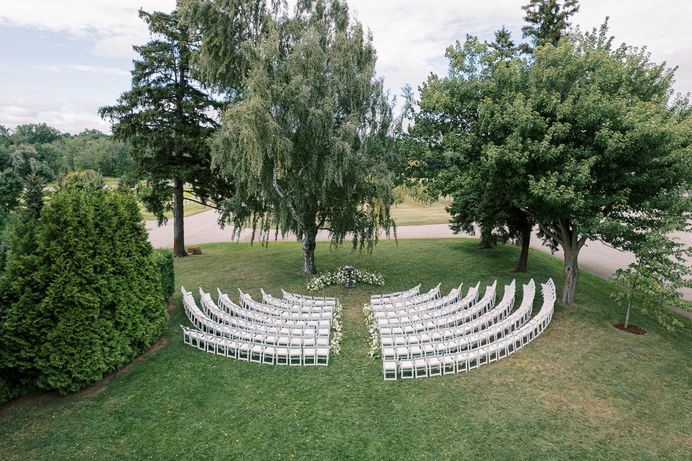 Outdoor wedding ceremony setup with white chairs arranged in a semi-circle on green lawn, surrounded by trees, perfect for a natural and elegant event.