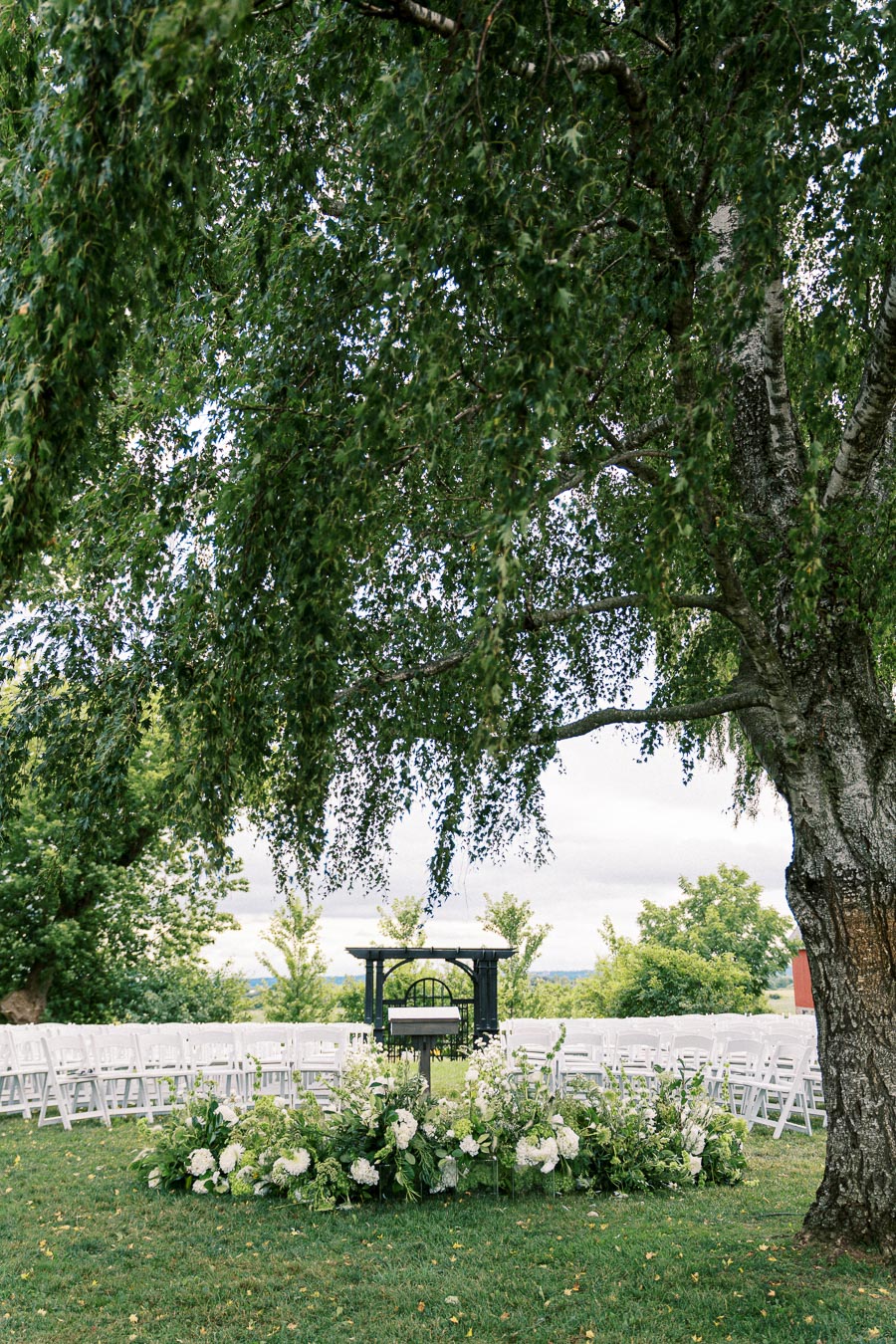 Outdoor wedding ceremony setup under a large tree with white chairs and floral arrangements surrounding an altar.