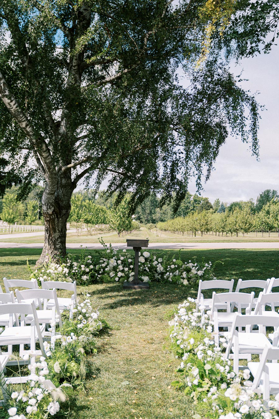 Outdoor wedding ceremony setup with white chairs and floral arrangements under a large tree in a scenic garden setting.