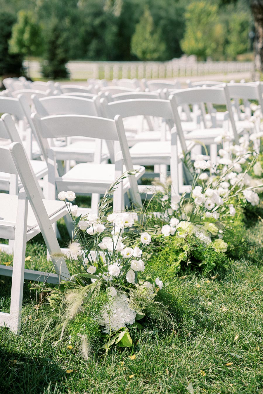 Outdoor wedding setup with white folding chairs and lush white floral arrangements on green grass under a sunny sky.
