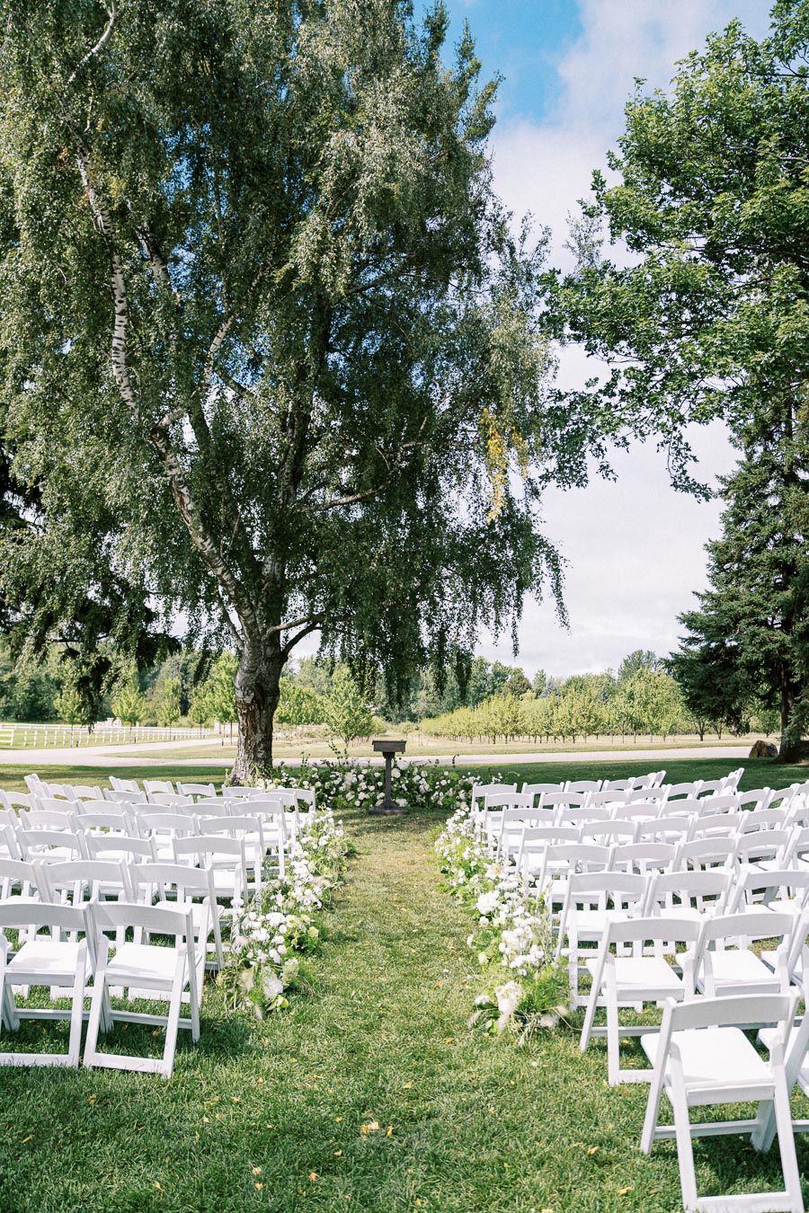 Outdoor wedding ceremony setup featuring white chairs and floral arrangements on a grassy aisle, under a large tree in a serene garden setting.