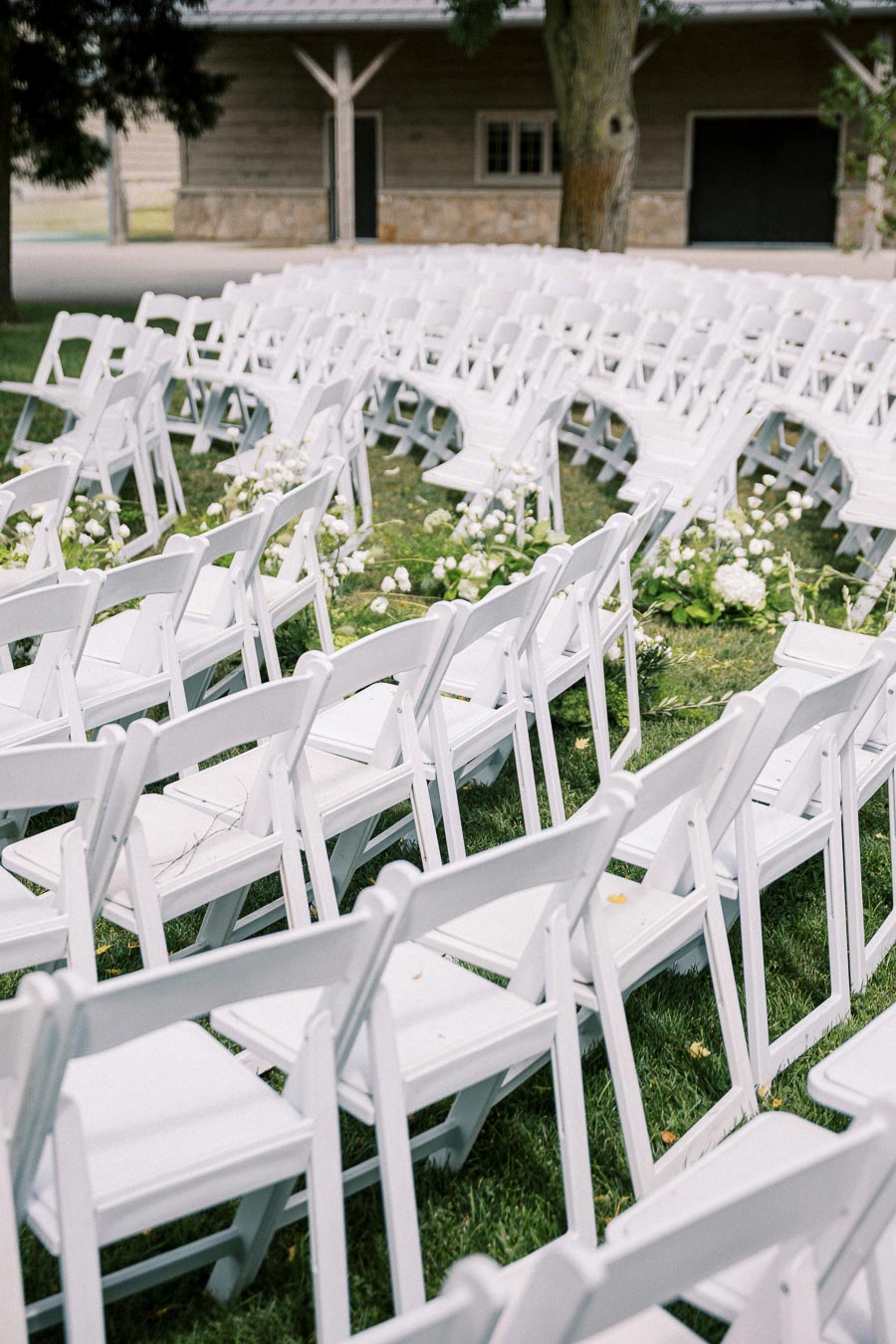 Outdoor wedding ceremony setup with white folding chairs arranged in rows on a grassy lawn, adorned with white flowers and a rustic wooden structure in the background.