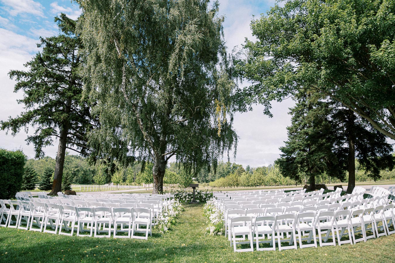 Outdoor wedding ceremony setup in a scenic garden, featuring rows of white chairs arranged between tall, lush trees under a clear blue sky.