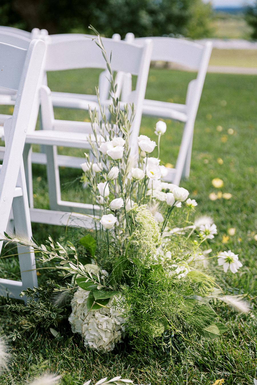 Outdoor wedding ceremony setup featuring white folding chairs and elegant floral arrangements with white flowers and green foliage on a lush grassy lawn.