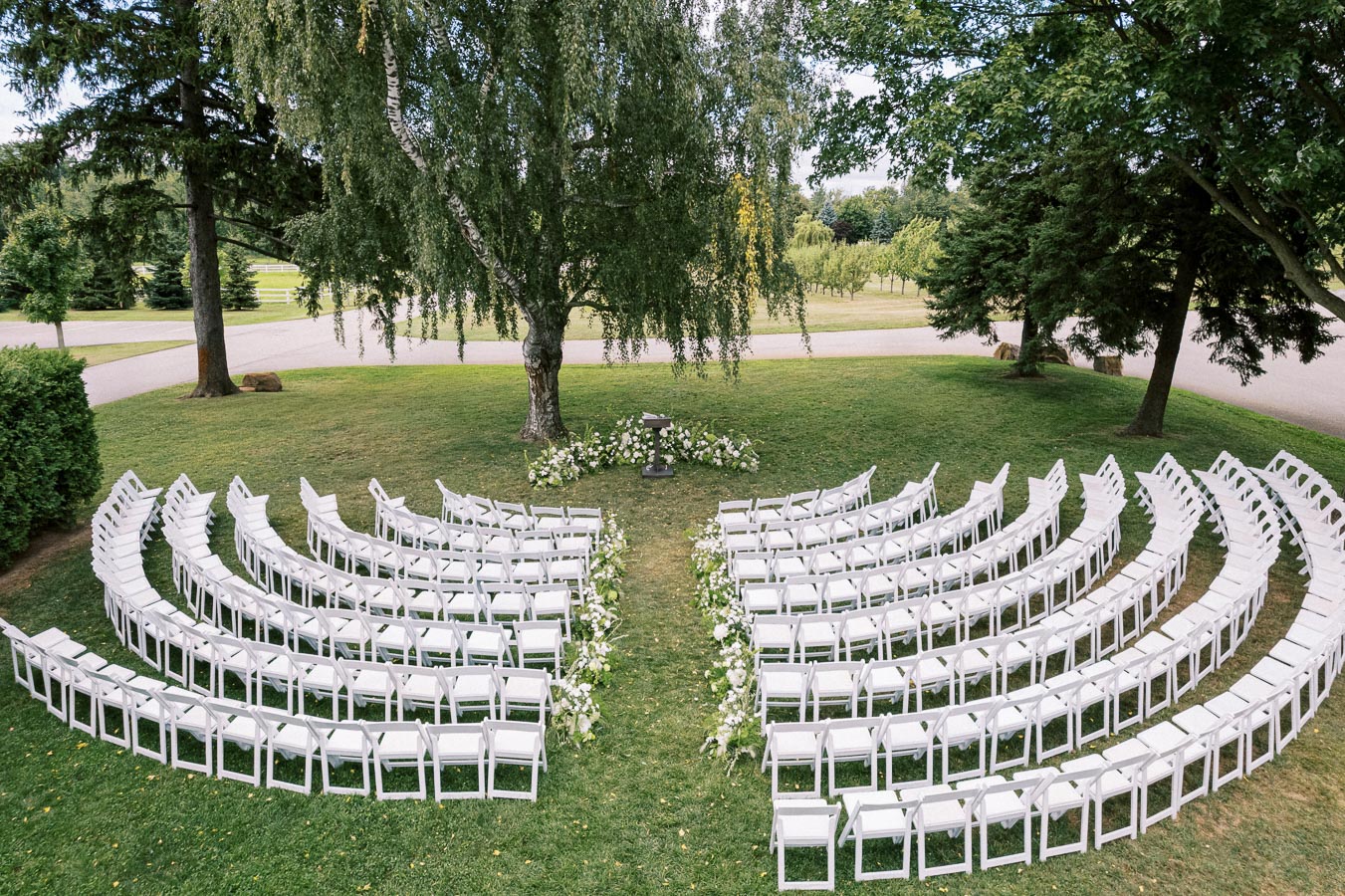 Outdoor wedding ceremony setup with white chairs arranged in a semi-circle on a lawn, surrounded by green trees and floral decorations under a clear sky.