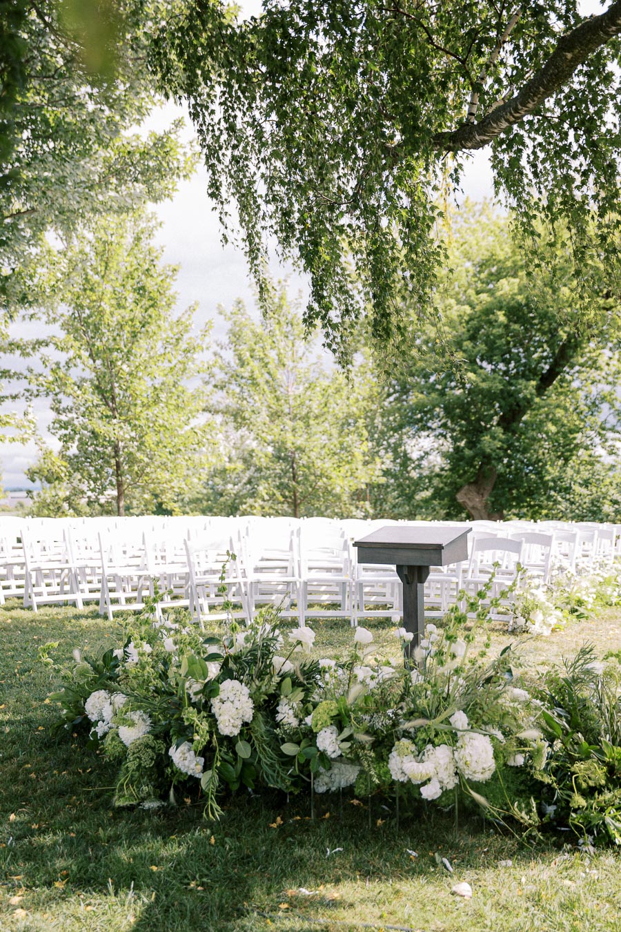 Outdoor wedding ceremony setup with white chairs, lush greenery, and a floral arrangement, under a canopy of leafy trees.