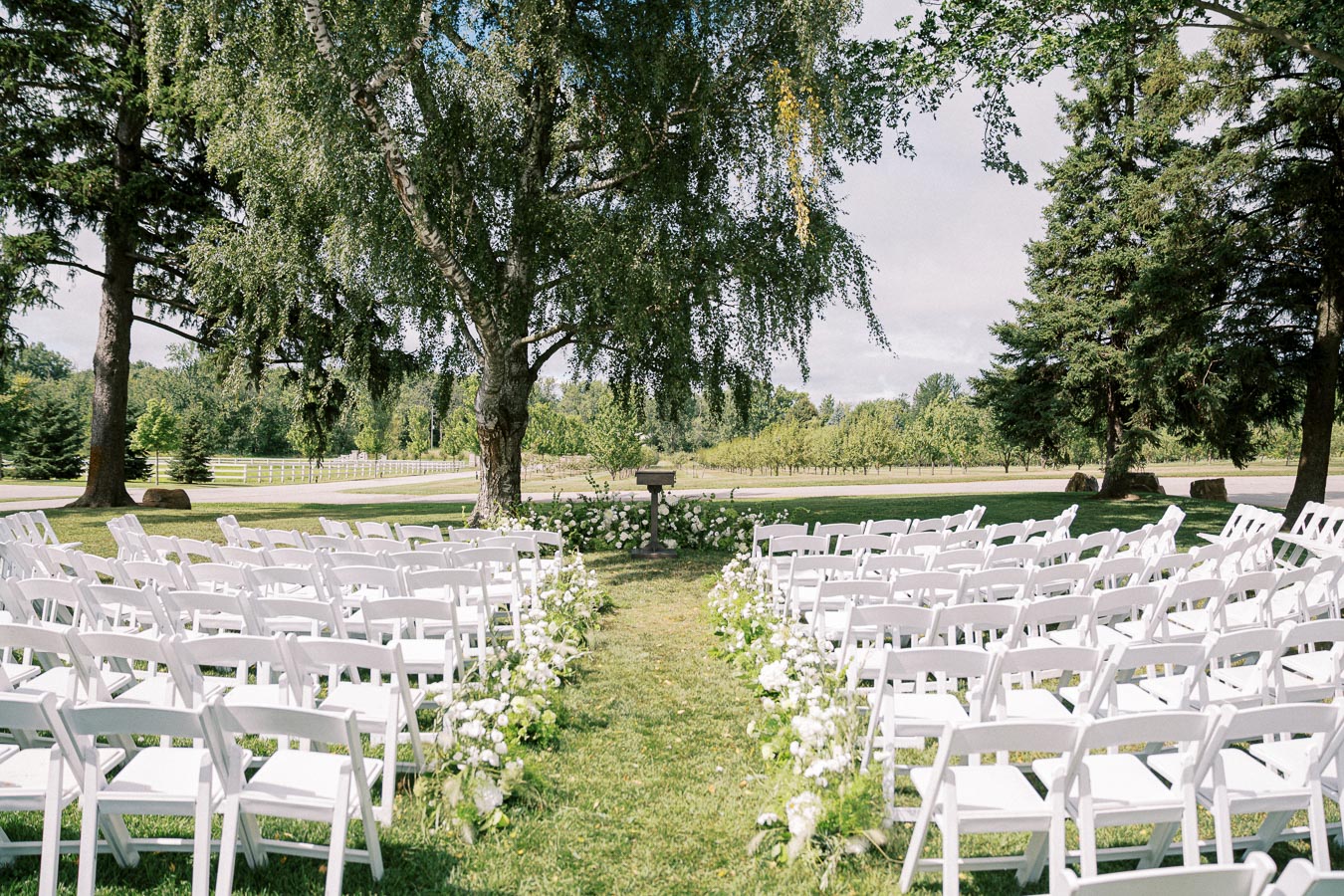 Outdoor wedding ceremony setup with white chairs arranged in rows on a green lawn, flanked by lush trees and floral decorations under a clear sky.