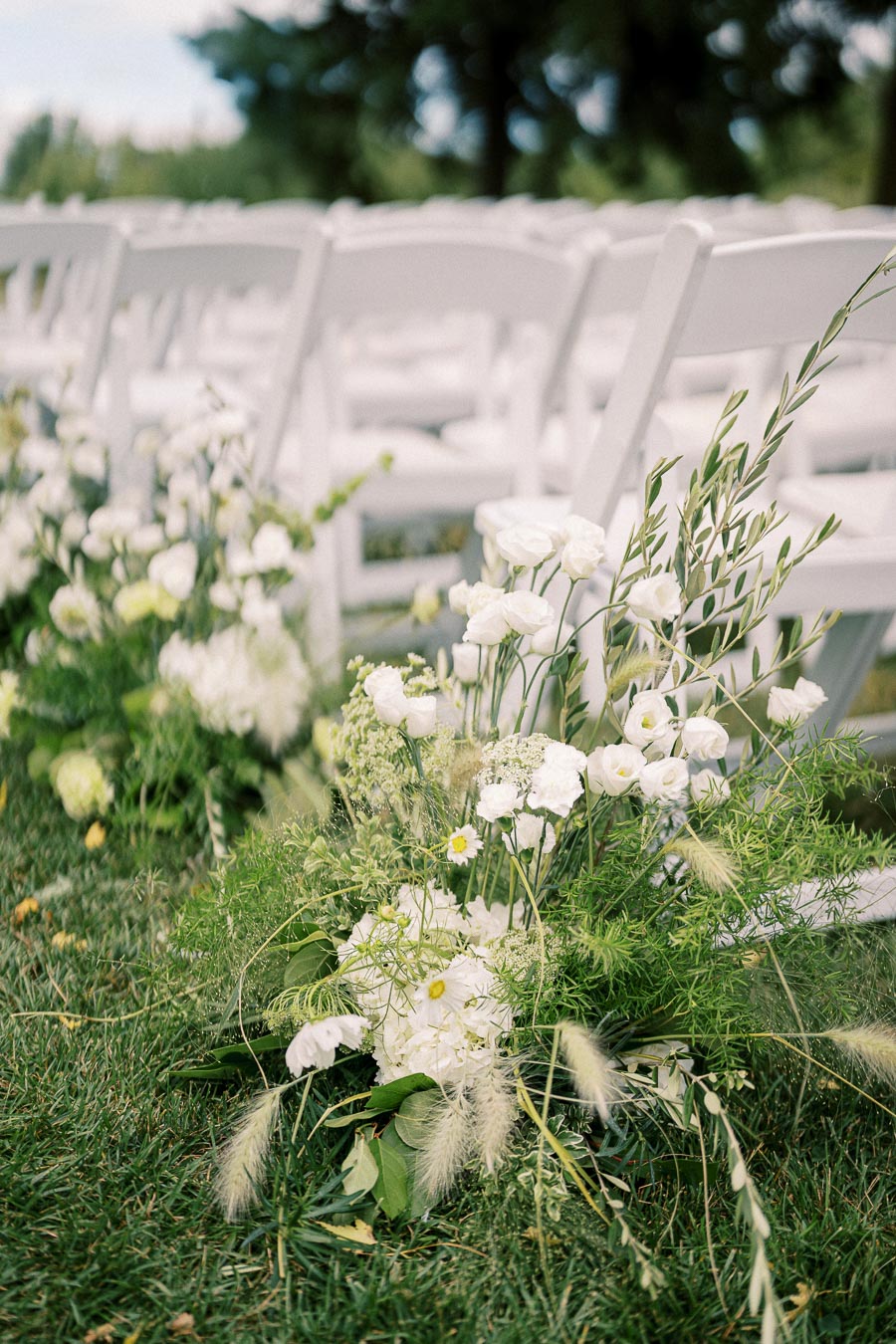 Elegant outdoor wedding ceremony setup with white chairs and lush floral arrangements, including white flowers and greenery, on a manicured lawn.