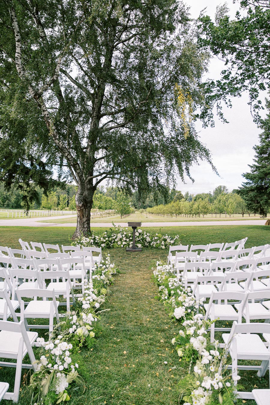 Outdoor wedding ceremony setup with white chairs and floral decorations on a grassy aisle, under a large tree in a scenic garden setting.