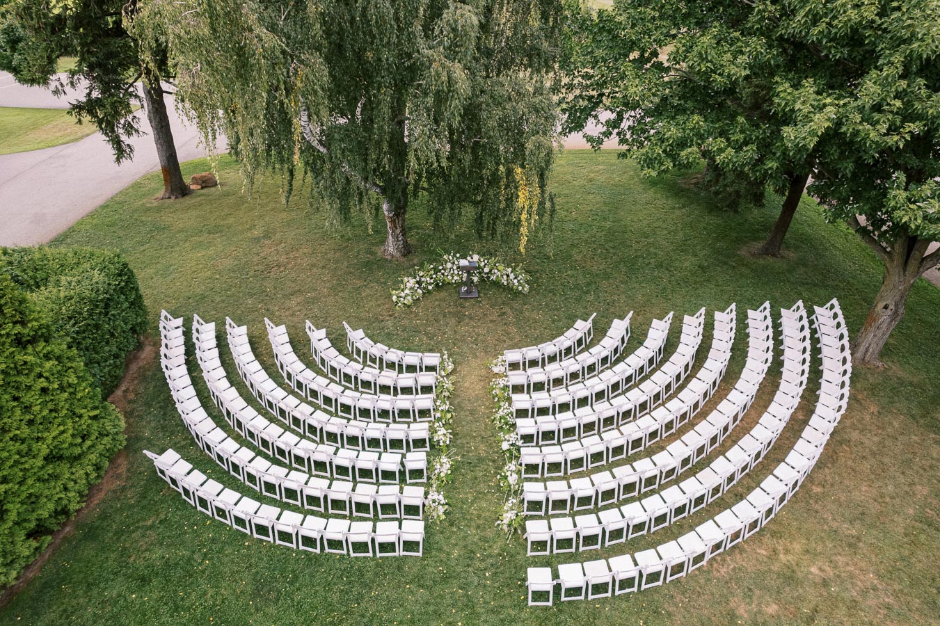 Aerial view of an outdoor wedding ceremony setup featuring white chairs arranged in a semi-circle under a large tree, surrounded by lush greenery.