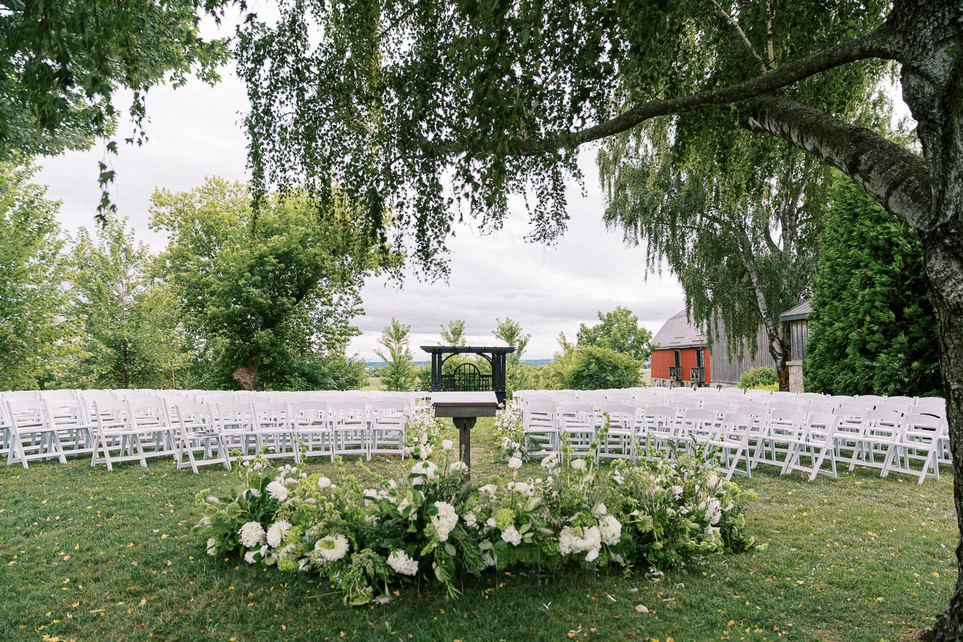 Outdoor wedding ceremony setup with white chairs arranged on a grassy lawn, surrounded by lush greenery and trees, featuring a floral altar at the center.