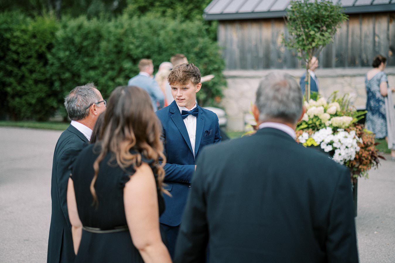 A young man in a blue suit and bow tie engaged in conversation with three elegantly dressed individuals at an outdoor event, with a background of greenery and a wooden building.