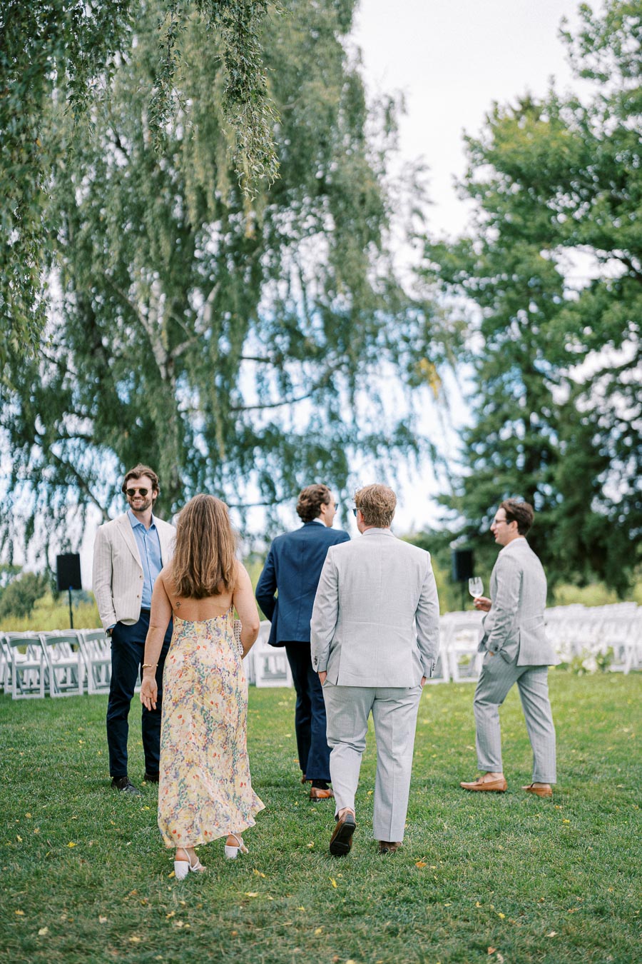 Group of elegantly dressed people walking through an outdoor garden setting, surrounded by greenery and white chairs, perfect for a summer wedding or garden party.