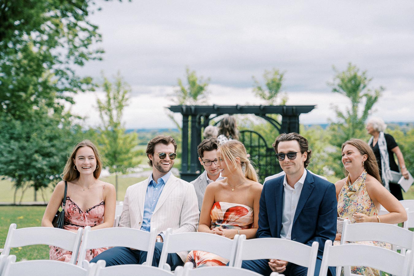 A group of young adults, dressed in stylish attire, sitting outdoors on white chairs, smiling and chatting against a backdrop of trees and a cloudy sky, capturing a joyful and relaxed moment.