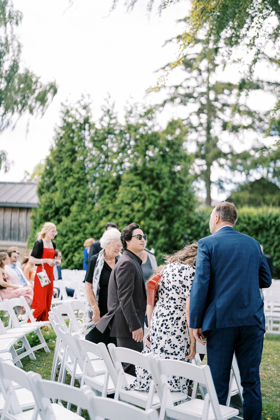 A group of well-dressed guests socializing at an outdoor wedding ceremony. White chairs are arranged on a grassy area, surrounded by lush greenery and trees in the background. The scene captures the joyful ambiance of a sunny day celebration.