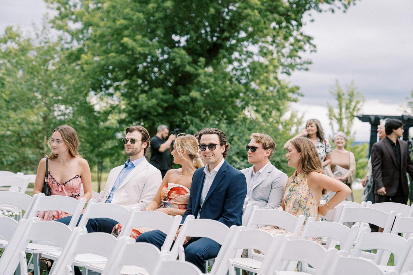 Group of young adults sitting in white chairs at an outdoor event, dressed in summer attire and sunglasses, with a lush green background.