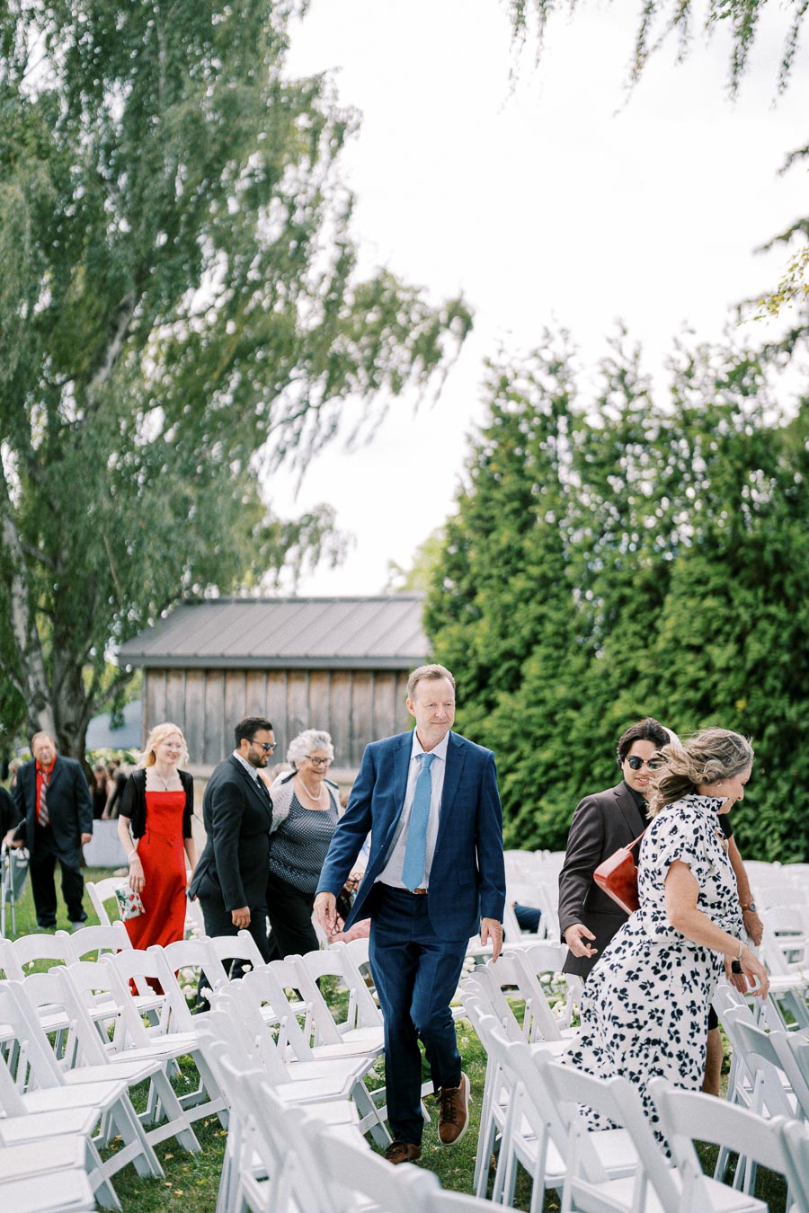 A group of well-dressed guests walking through a garden setting with white chairs, attending an outdoor event. Tall trees and a rustic wooden building are in the background, creating an elegant atmosphere.