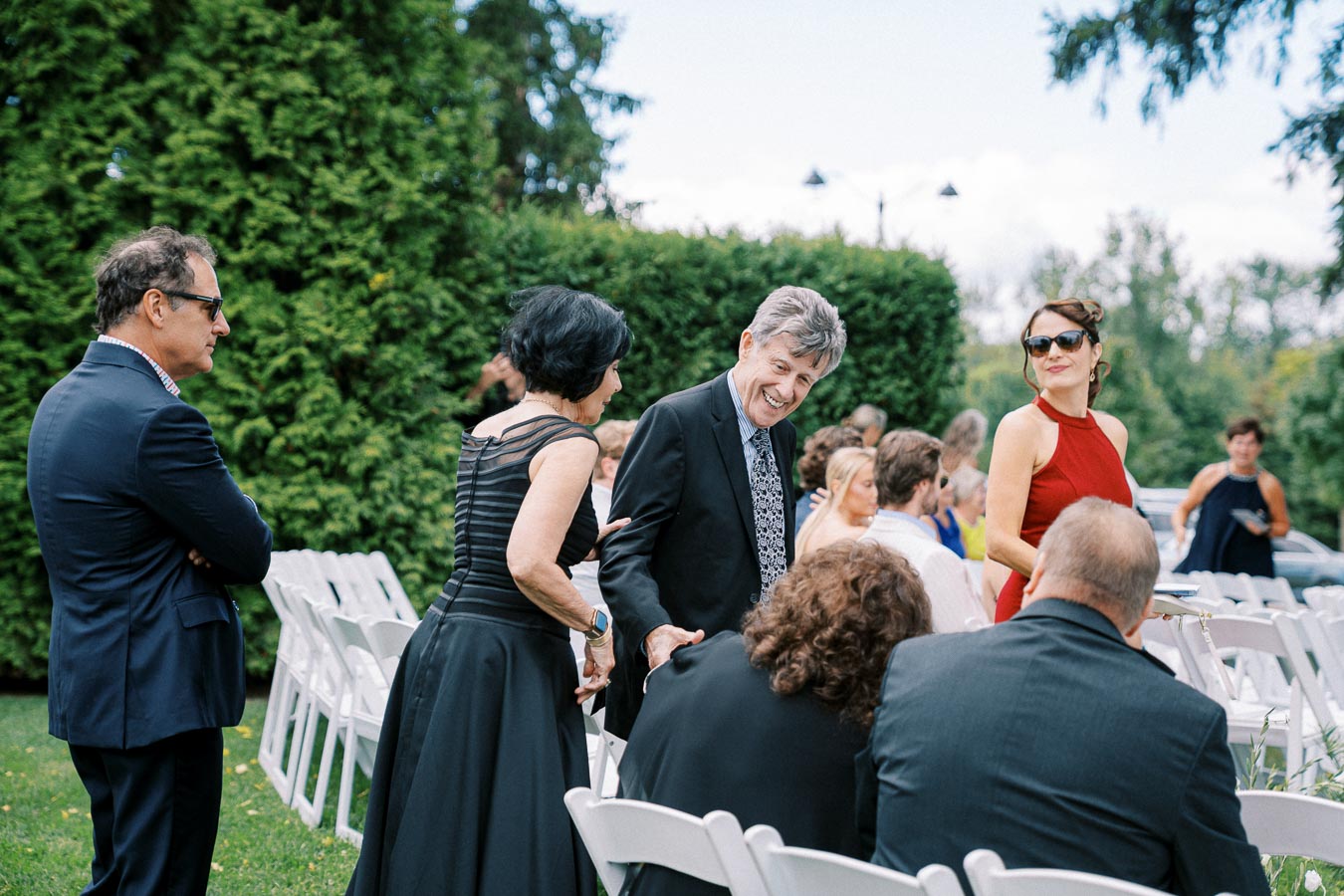 A group of elegantly dressed people socializing at an outdoor event, surrounded by lush greenery, with white chairs arranged on a grassy lawn.