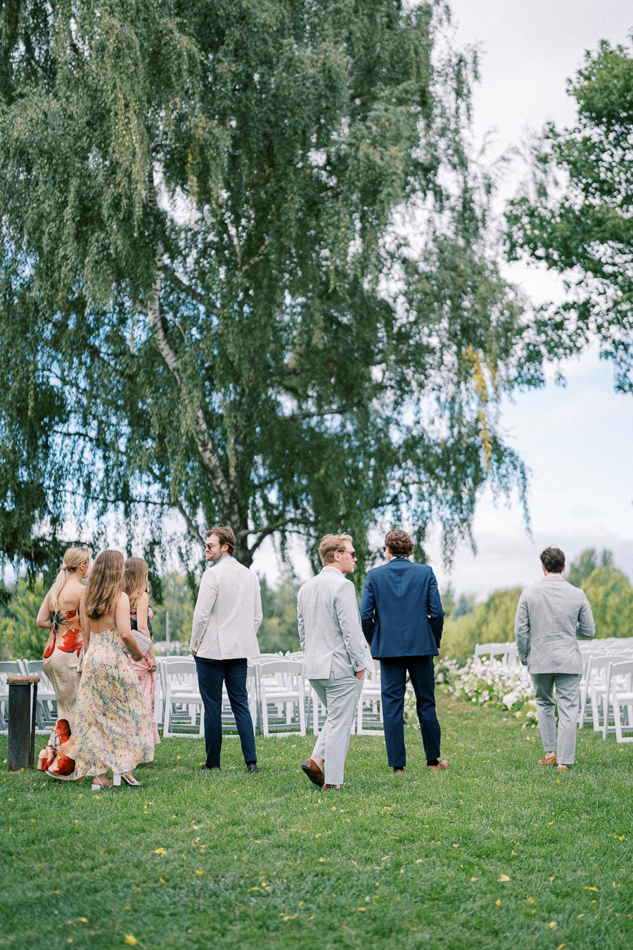 Group of elegantly dressed guests walking through a scenic outdoor wedding venue with white chairs and lush greenery.