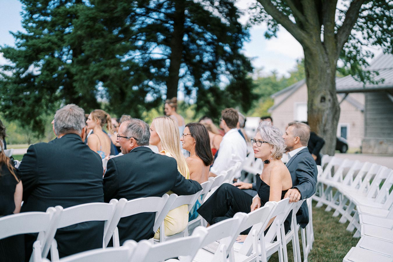 A group of guests seated on white chairs outdoors at a wedding ceremony, under the shade of a large tree, with a house visible in the background, dressed in formal attire and engaged in conversation.