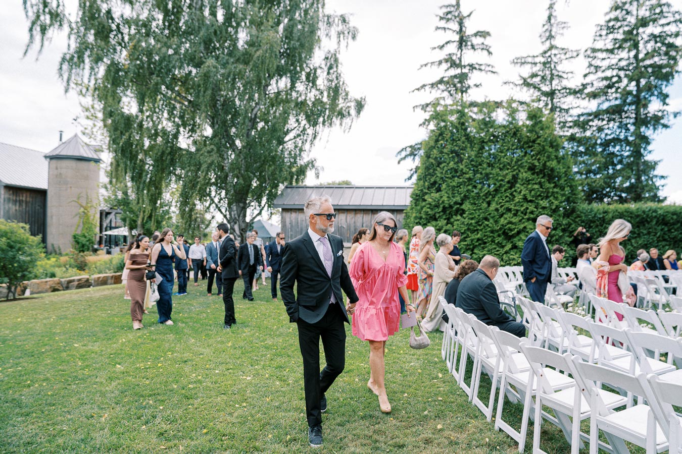 Guests arriving at an outdoor wedding ceremony set in a picturesque garden with lush trees and rustic barns, wearing formal attire and seated on white chairs.