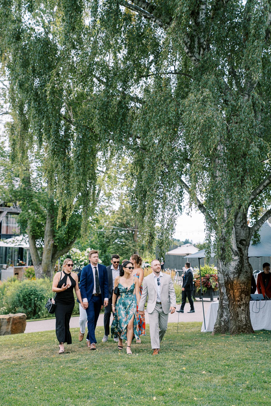 A group of well-dressed guests walking outdoors at a garden wedding, surrounded by lush greenery and trees.