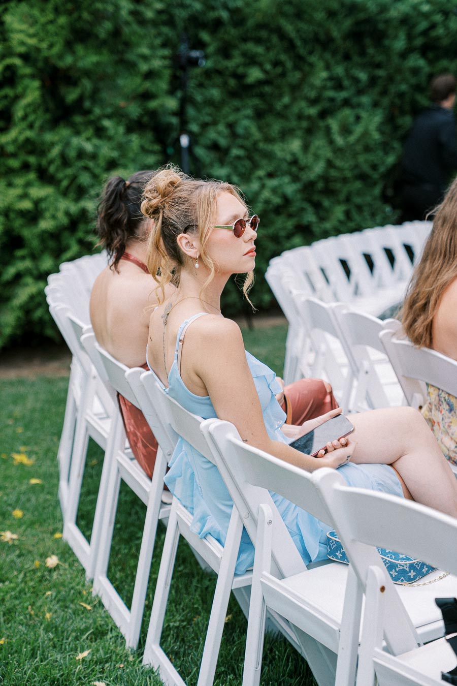 A woman in a blue dress and sunglasses sitting on white folding chairs in an outdoor setting, surrounded by greenery, holding a smartphone.