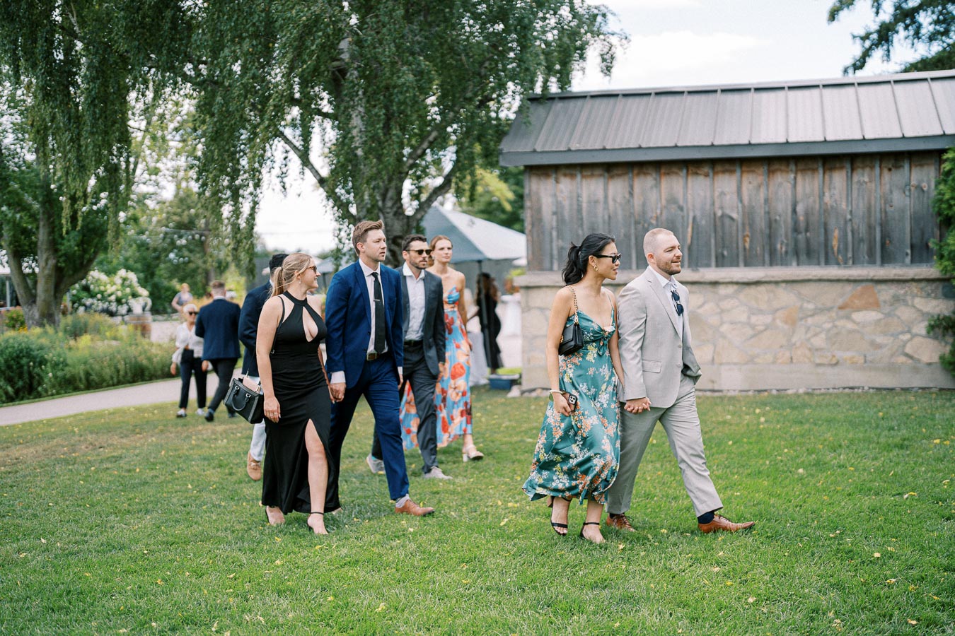 A group of well-dressed people walking on a grassy lawn, with some wearing suits and elegant dresses, in front of a rustic wooden building and trees.