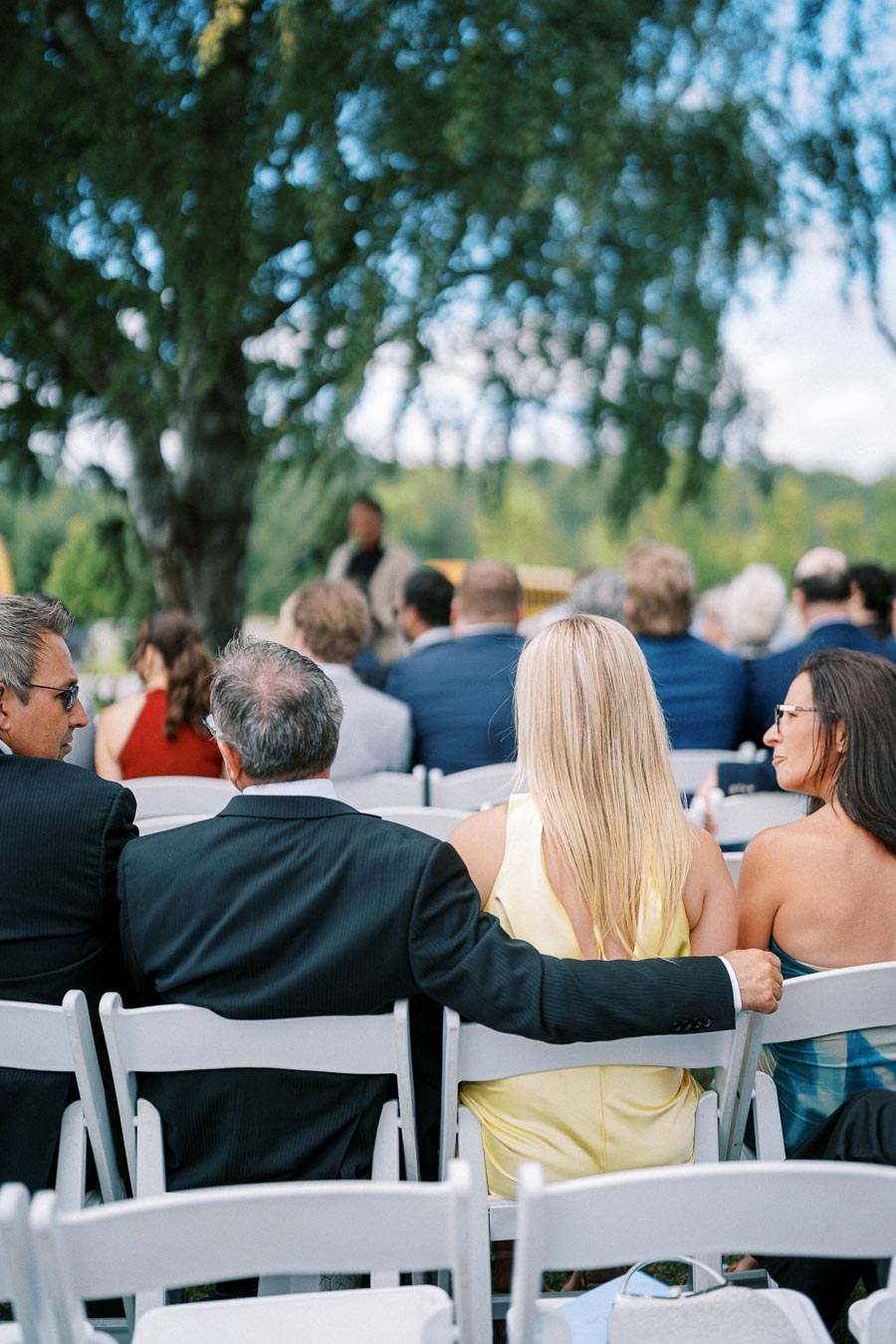 Guests seated outdoors at a formal event, with a focus on a man in a suit and two women dressed in elegant attire, surrounded by a gathering under lush green trees.