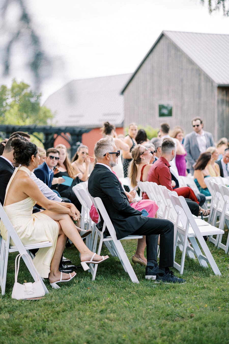 Outdoor wedding ceremony with guests seated in white chairs, dressed in formal attire, set against a rustic barn backdrop.