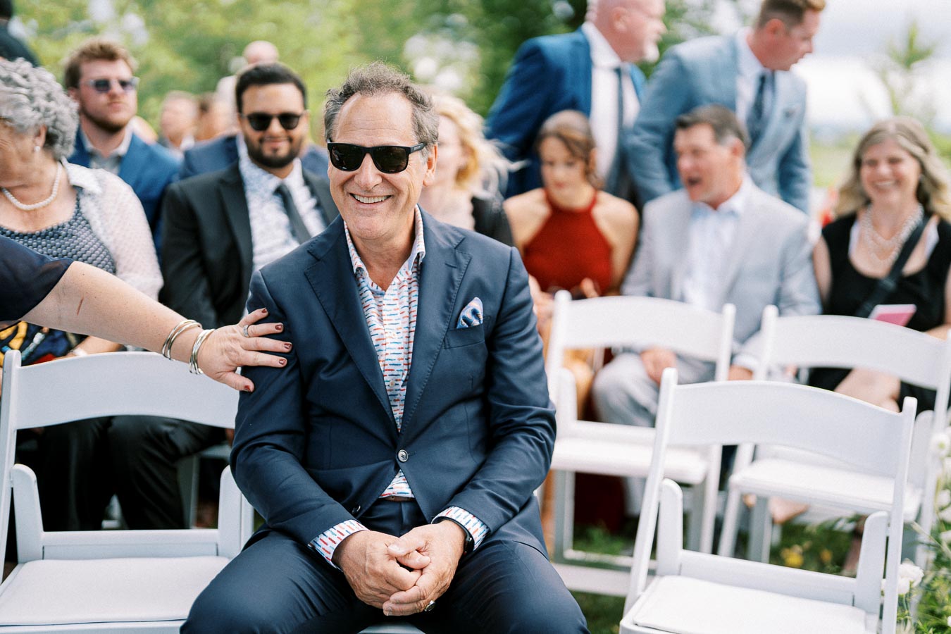 Smiling man in sunglasses sitting on a white chair at an outdoor event, surrounded by well-dressed people on a sunny day.
