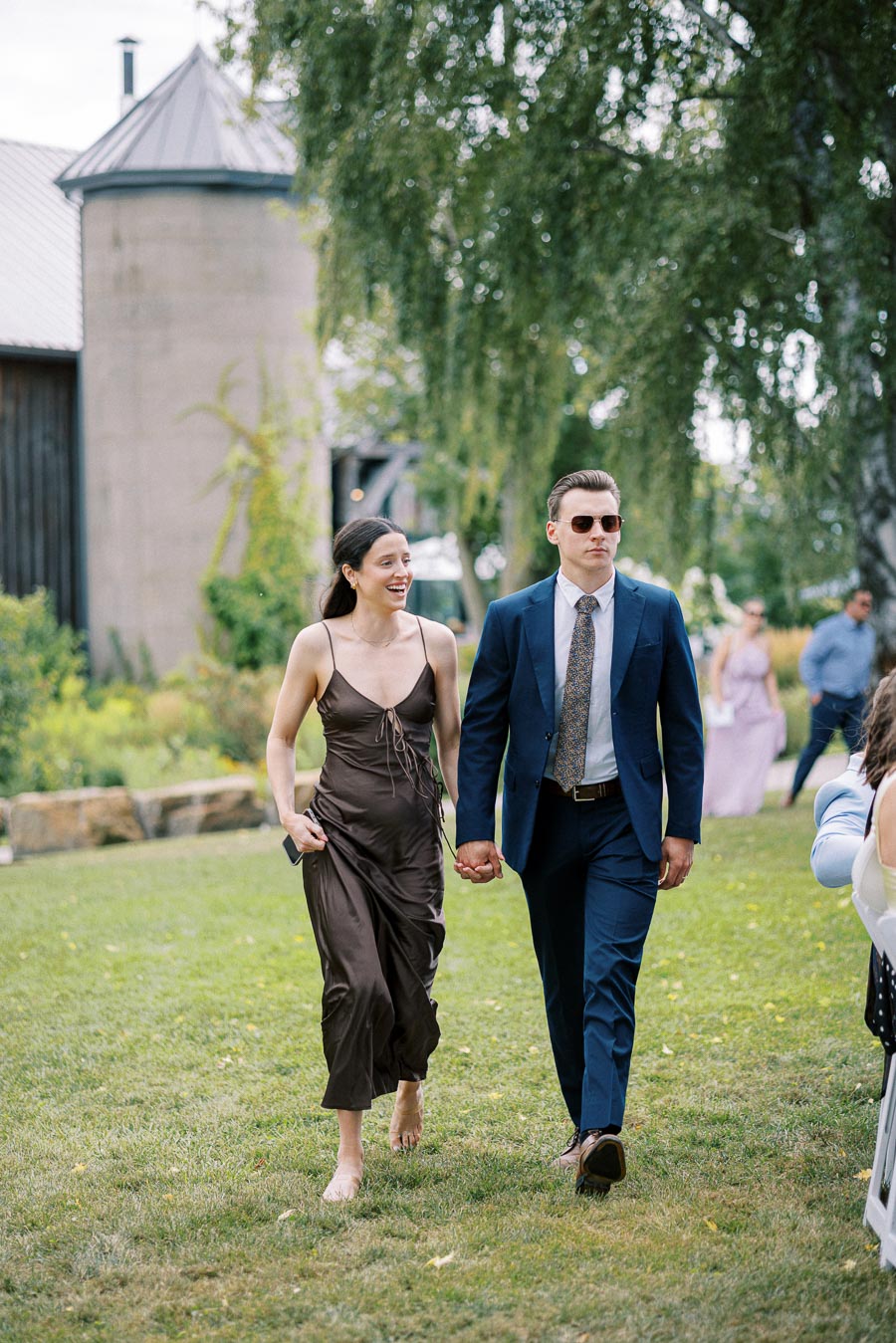 A couple walks hand-in-hand on a lawn at an outdoor event, with the man in a blue suit and sunglasses and the woman in a brown dress, against a backdrop of greenery and rustic buildings.