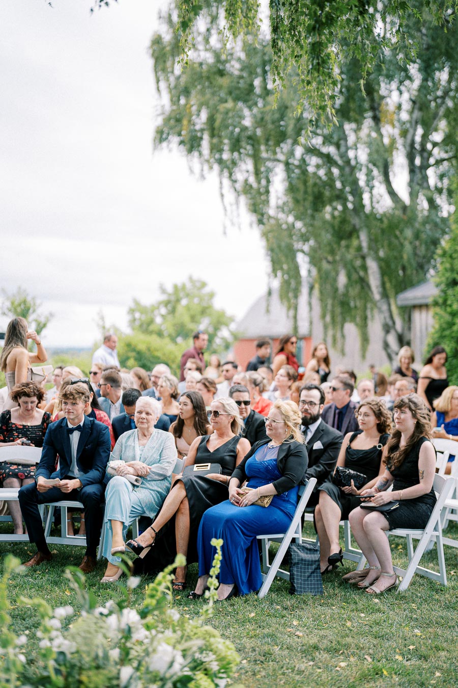Outdoor wedding ceremony with guests seated on white chairs, dressed in formal attire, surrounded by lush greenery and flowers under a clear sky.