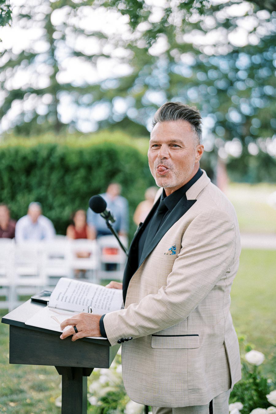 A man in a beige suit making a humorous expression while speaking at an outdoor event, microphone and notes on a podium.