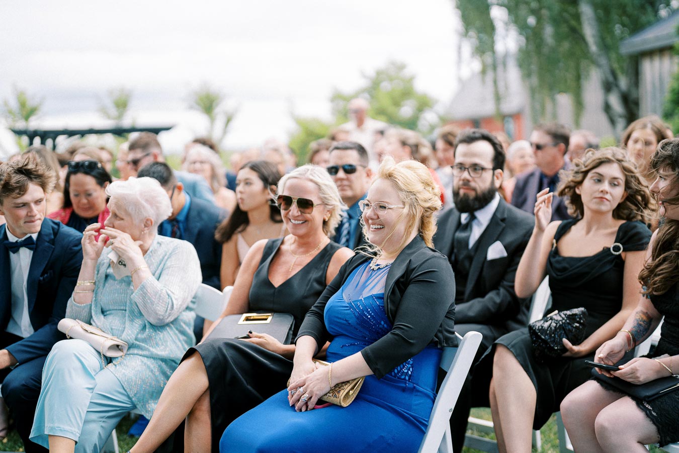 Outdoor wedding ceremony with guests seated in chairs, dressed in formal attire, and smiling on a sunny day.