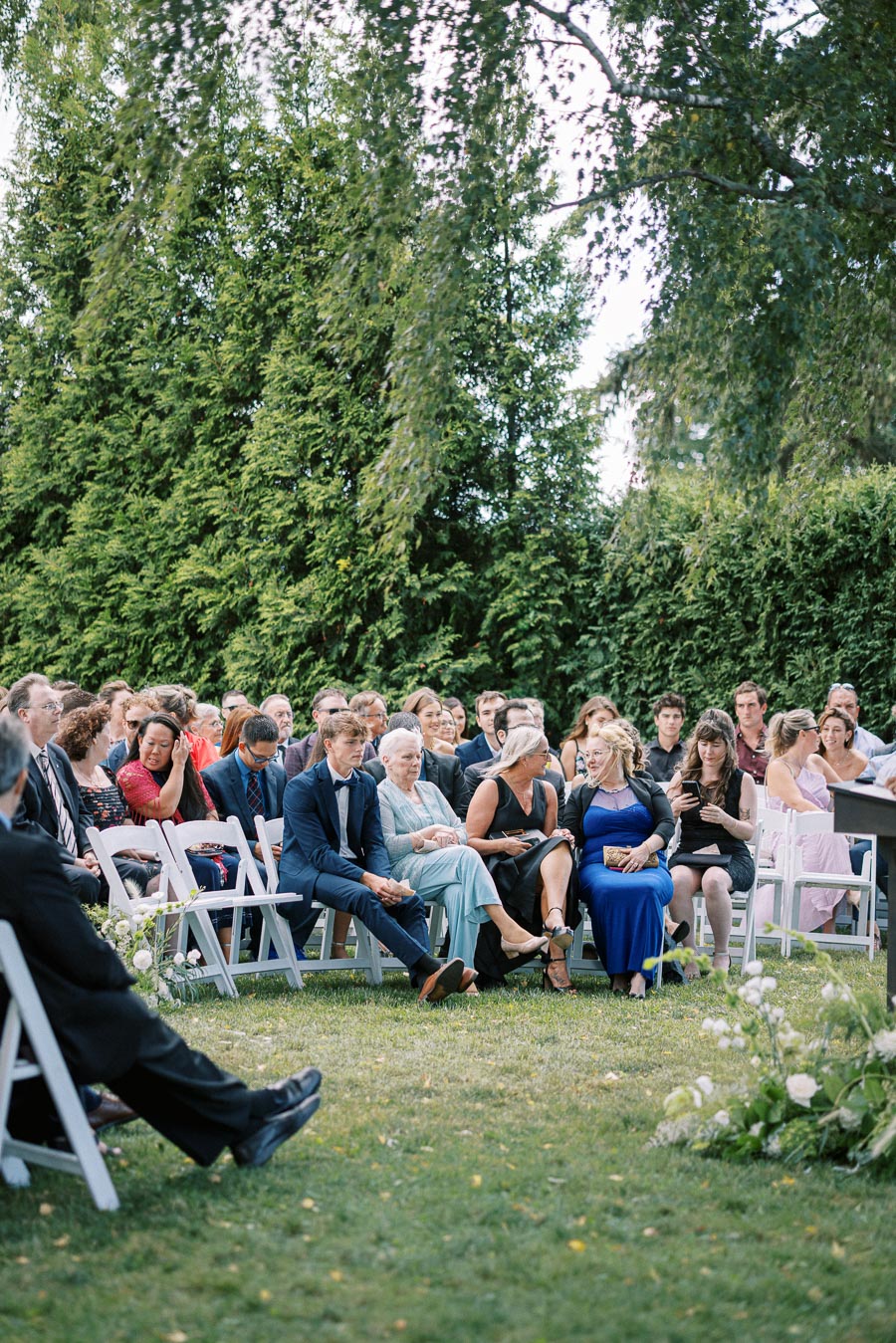 Outdoor wedding ceremony with guests seated on white chairs, surrounded by lush greenery, engaged in conversation and awaiting the event.