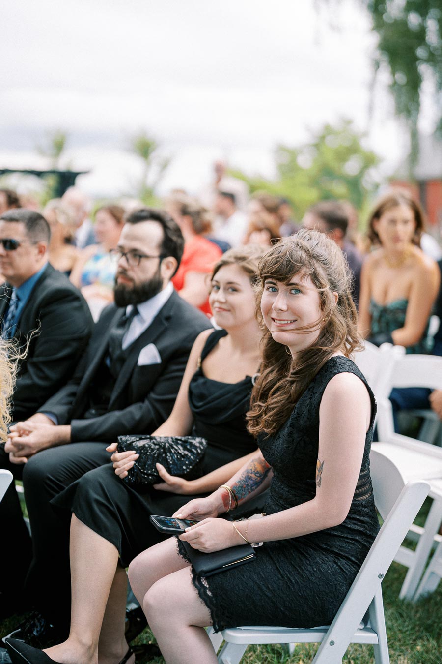 A group of people seated outdoors at a social event, with a focus on a smiling woman in a black dress holding a smartphone. The attendees are dressed in formal attire, and the background is slightly blurred, suggesting a lively gathering.