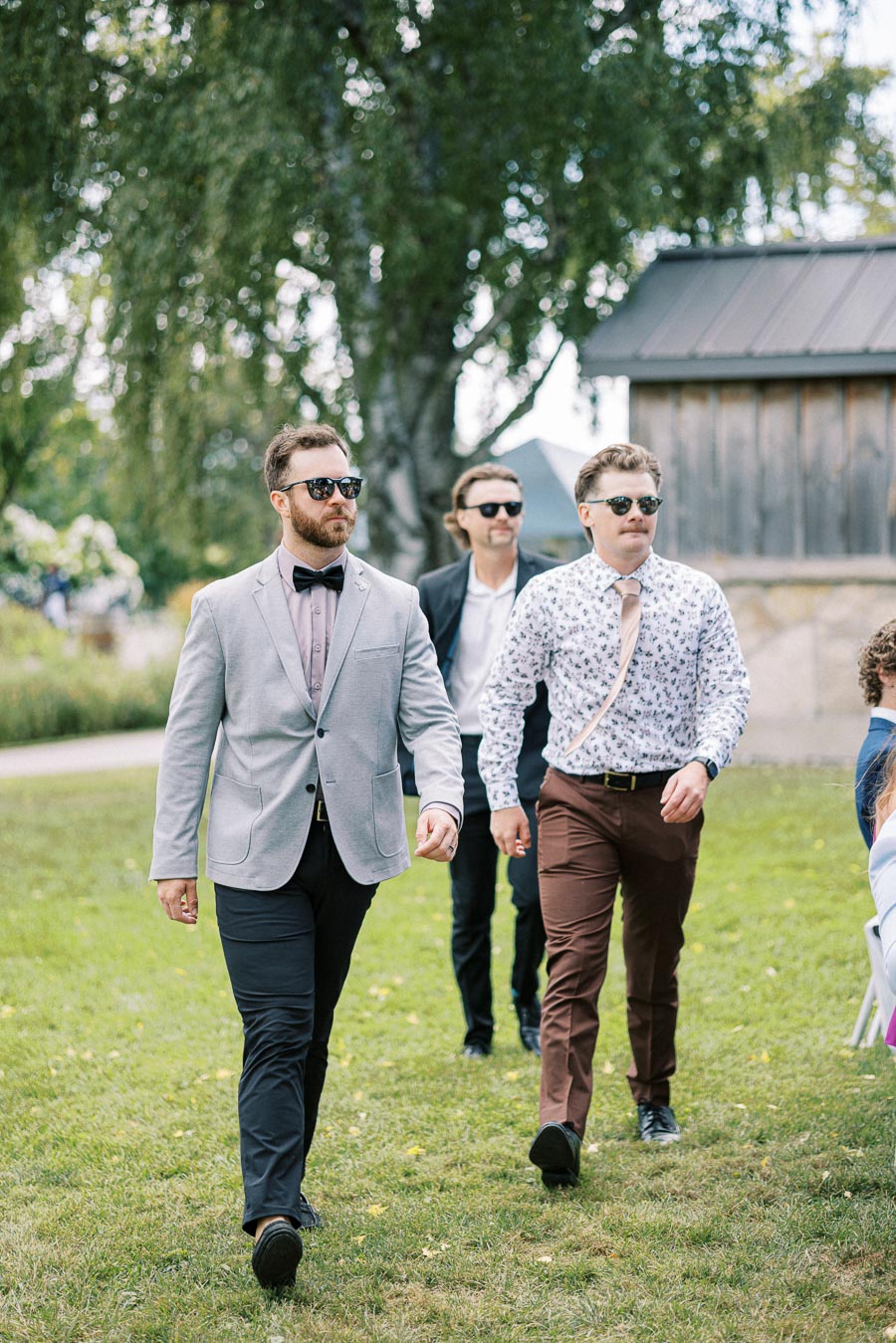 Three men wearing stylish outfits and sunglasses walking confidently outdoors on a grassy area, with trees and a wooden cabin in the background, suggesting a casual event or gathering.