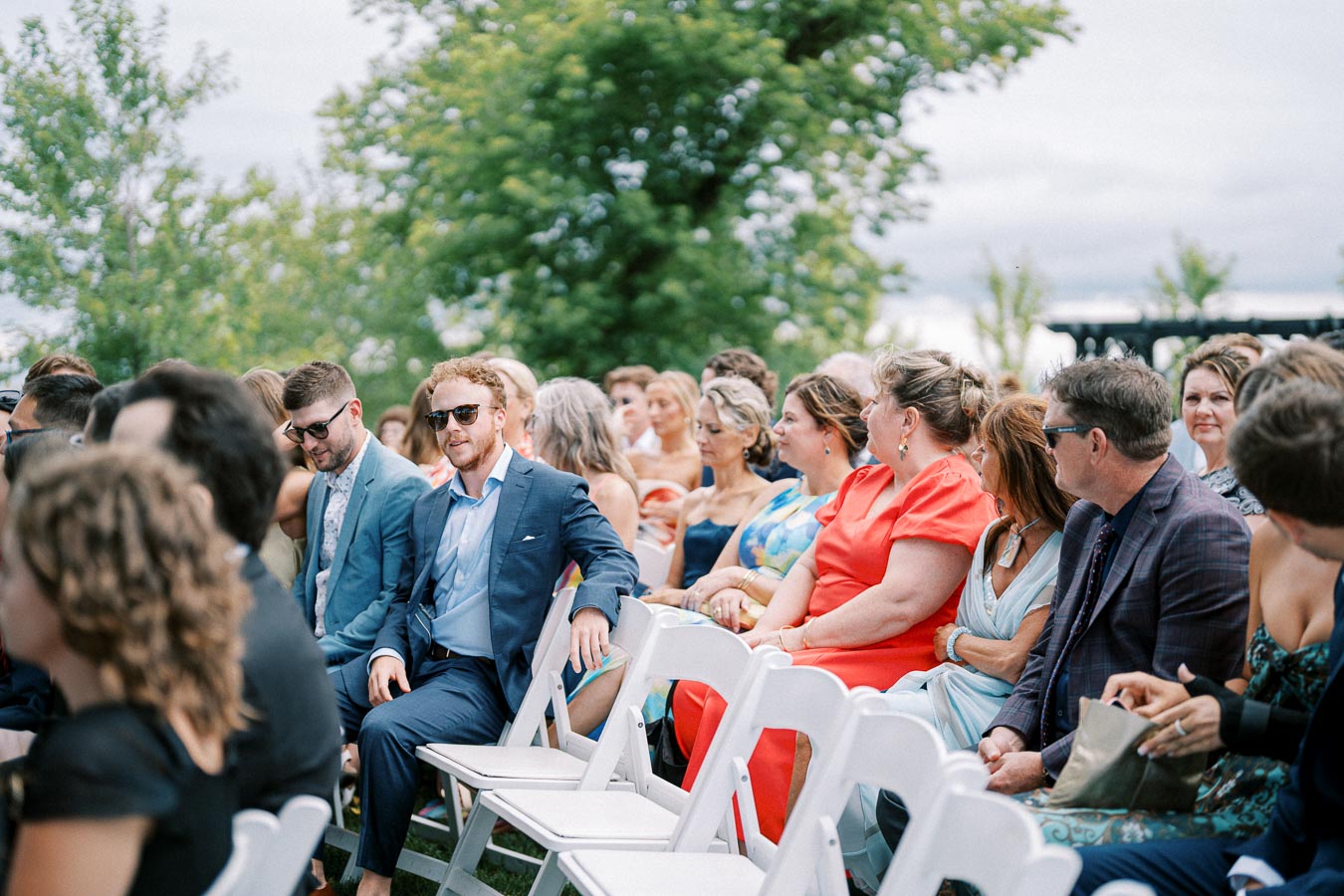 Outdoor wedding ceremony guests seated on white chairs, wearing colorful formal attire and sunglasses, in a lush green garden setting with clear skies.