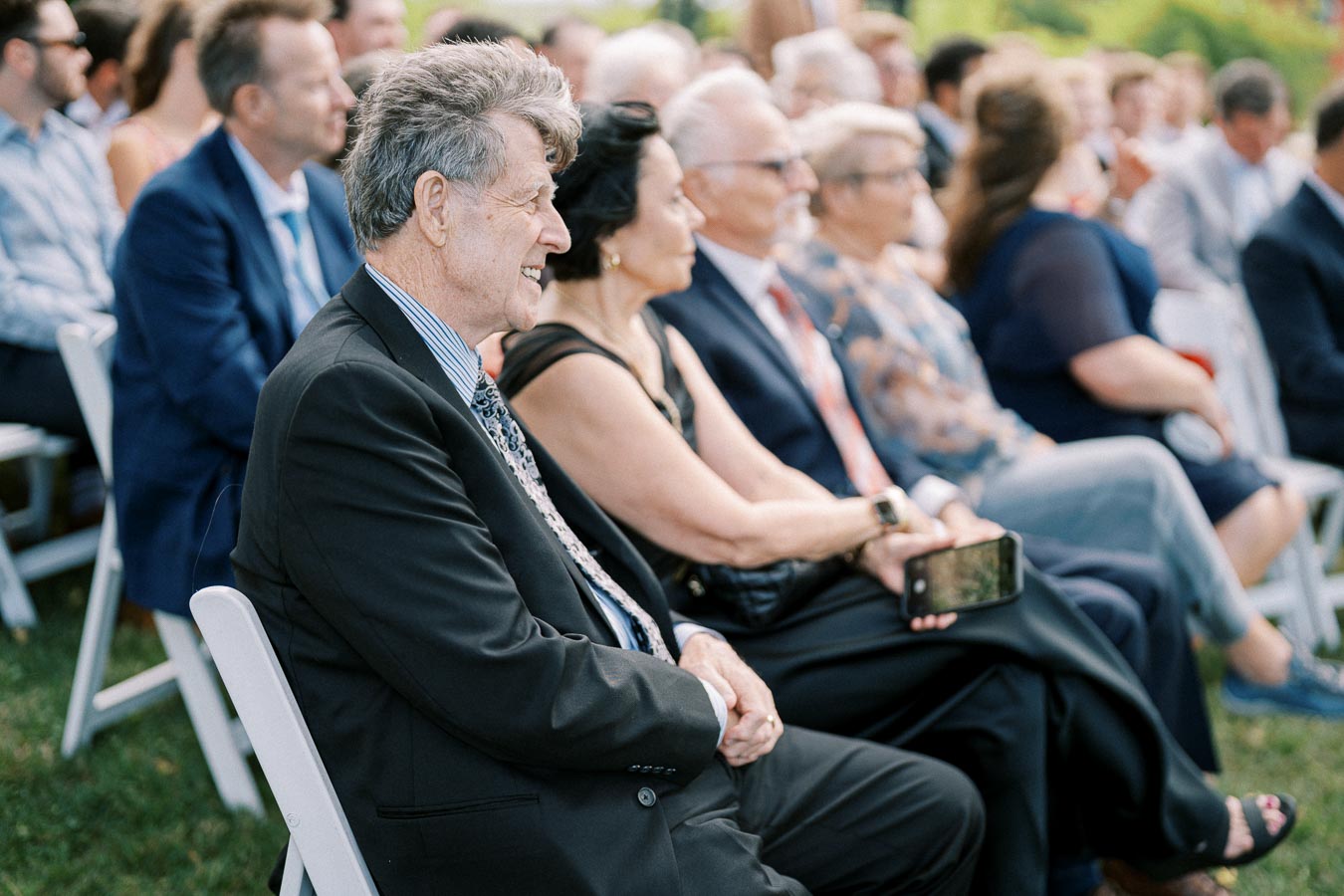 Elderly man in a suit smiling during an outdoor event, seated among a diverse group of formally dressed attendees, capturing a moment of joy and engagement.