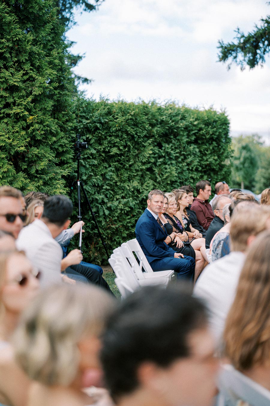 Outdoor wedding ceremony with guests seated on white chairs, surrounded by lush green hedges on a sunny day.