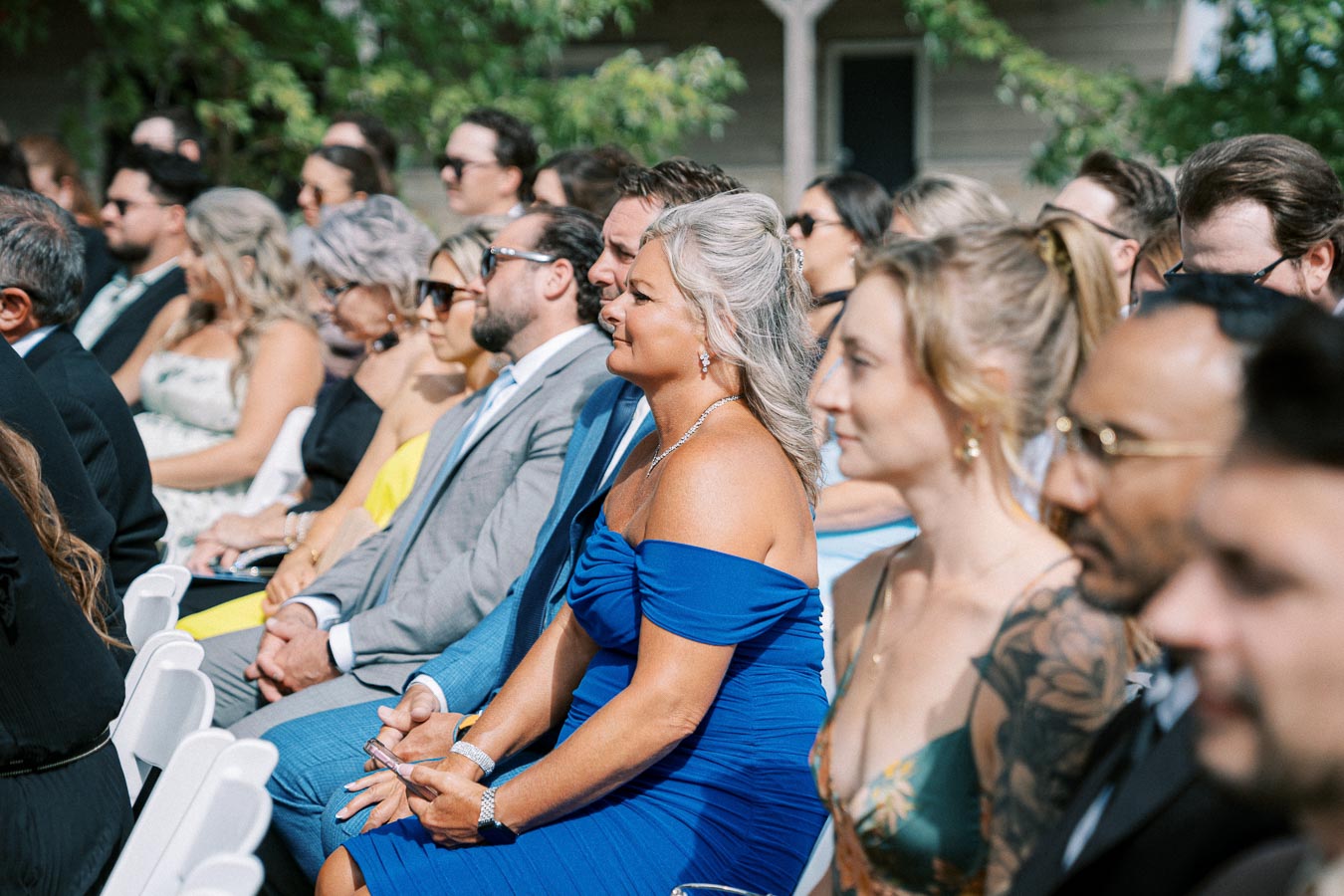 Audience at an outdoor wedding, dressed in formal attire, seated in white chairs, attentively watching the ceremony under sunny skies with green foliage in the background.