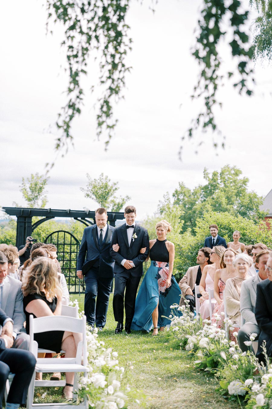 Outdoor wedding ceremony with guests seated on white chairs, groom walking down the aisle with escorts, surrounded by greenery and floral decorations.