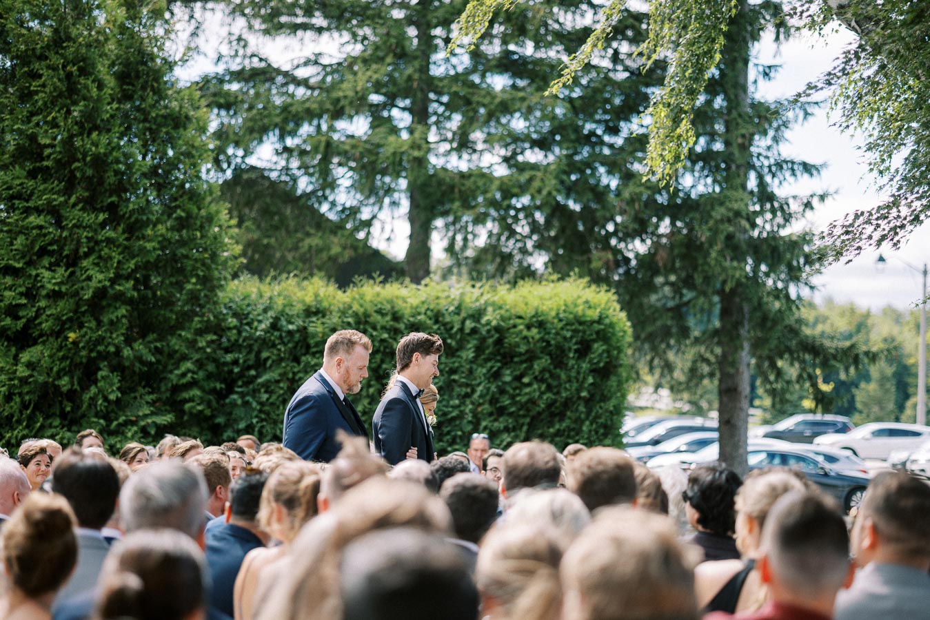 Two men in suits walking down an outdoor aisle at a wedding ceremony, surrounded by seated guests and lush greenery.