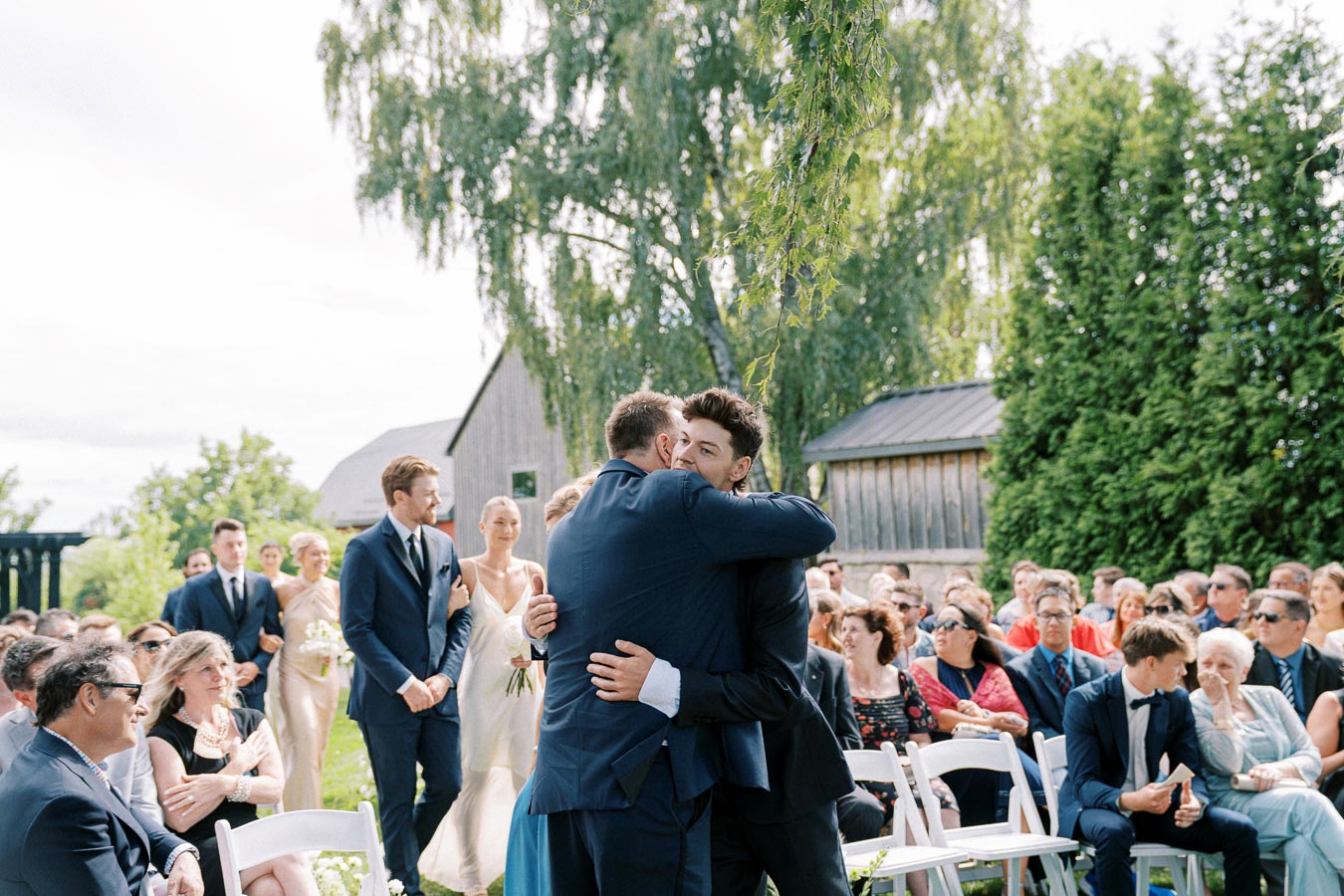 Outdoor wedding ceremony with a heartfelt embrace between two men in suits, surrounded by seated guests and a natural, rustic backdrop.