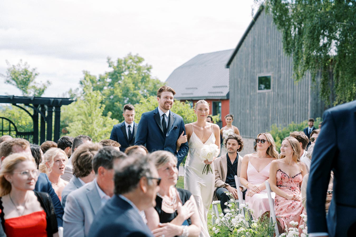 A joyful wedding procession with a smiling couple walking down an outdoor aisle. The groom is in a navy suit while the bride holds a bouquet of white roses. Guests in elegant attire are seated amidst a scenic greenery background, with a rustic barn visible in the distance.