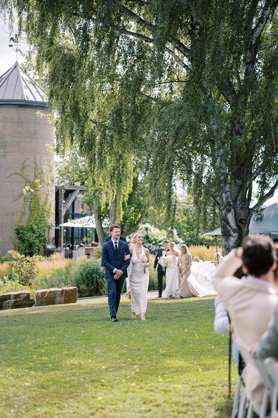 A couple walks down a grassy outdoor aisle during a wedding ceremony, surrounded by lush greenery and a large tree. Guests are seated, capturing the moment in a beautiful garden setting.