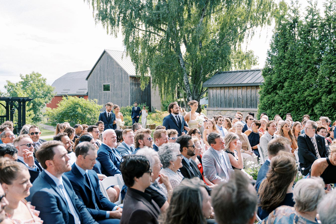 Outdoor wedding ceremony with guests seated, surrounded by lush greenery and rustic wooden buildings.