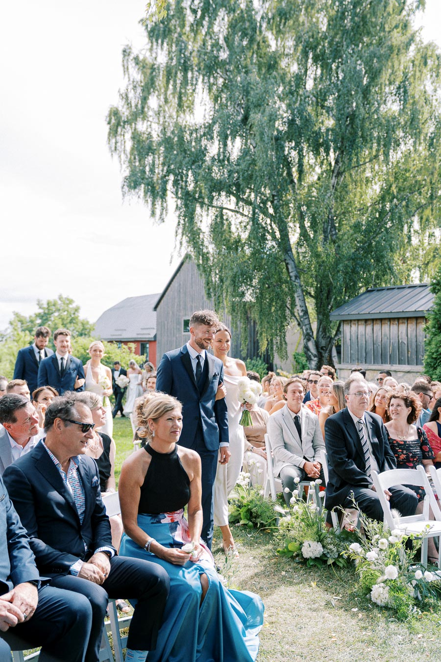 Outdoor wedding ceremony with a couple walking down the aisle, surrounded by seated guests, lush greenery, and rustic barns in the background.